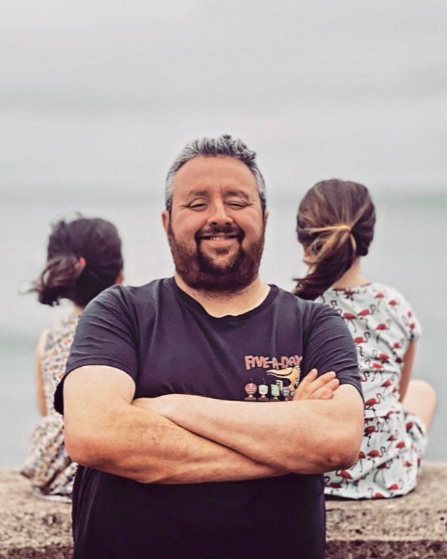 Jon Russell smiling with his arms folded, standing on a sea wall with two children sitting behind him looking out to sea.